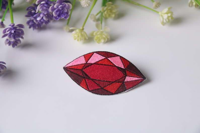 May include: An iron-on patch embroidered in red and pink, shaped like a gemstone. The patch is detailed with black stitching and has a geometric design.