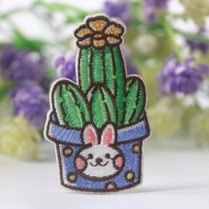 May include: Embroidered patch depicting a green cactus in a blue pot with a smiling bunny face. The patch has a brown border and is set against a blurred background of purple and green flowers. The cactus has brown flowers on top.