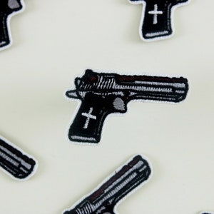 May include: Six black and white embroidered iron-on patches of a handgun with a cross on the handle.