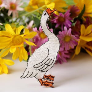 Puede incluir: Parche de ganso blanco bordado con pico y patas naranjas. El ganso está de pie sobre una superficie blanca con un fondo borroso de flores rosas y amarillas.