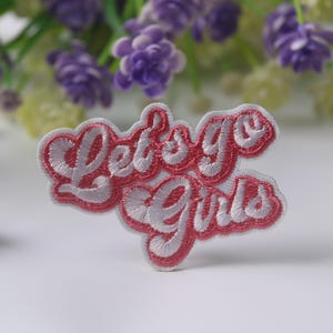 Puede incluir: Parche bordado con la frase "Let's Go Girls" en fuente cursiva. El parche presenta un diseño bordado en blanco con un contorno rosa. El parche está sobre una superficie blanca con flores moradas y amarillas en el fondo.