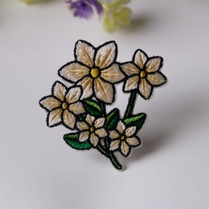 May include: A white embroidered flower patch with green leaves. The flowers have yellow centers and are arranged in a bouquet.