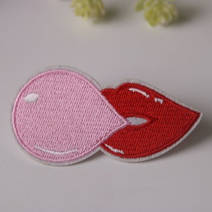 Könnte beinhalten: Ein gestickter Patch mit einem rosa Kaugummi und roten Lippen. Der Patch ist weiß mit rosa und roten Stickereien.