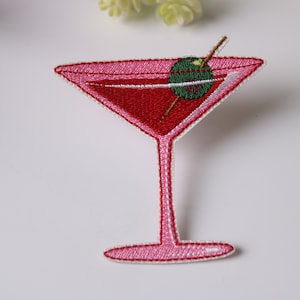 以下が含まれることがあります： 赤い飲み物とつまようじに刺さった緑のオリーブが入った、ピンク色の刺繍のアイロン接着パッチ。