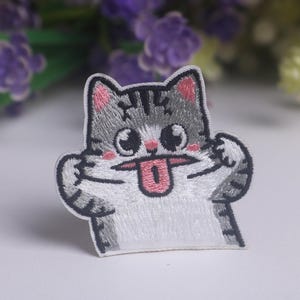 May include: An embroidered patch featuring a grey and white tabby cat with dark grey stripes, pink inner ears, and pink cheeks. The cat has large black eyes and is sticking out a pink tongue while holding its paws up near its face in a playful pose.