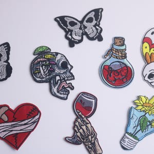 Op de afbeelding: Een verzameling geborduurde patches met diverse ontwerpen. Inbegrepen zijn schedelvormige vlinders, een schedel met paddenstoelen, een drankfles, een verbonden hart, een skelet hand met een wijnglas en een kapotte gloeilamp met een bloem.