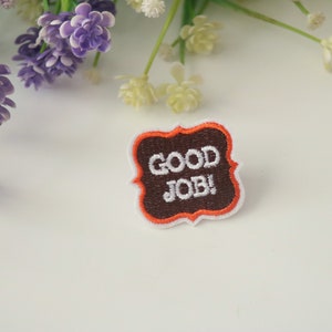 May include: A brown and white embroidered patch with an orange border. The patch is shaped like a banner and says "GOOD JOB!" in white text.