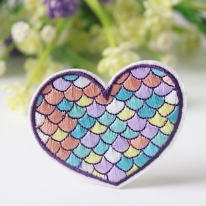 Può includere: Un cerotto ricamato a forma di cuore con un motivo a squame di sirena in tonalità di rosa, giallo, blu e viola. Le squame sono contornate da filo viola.