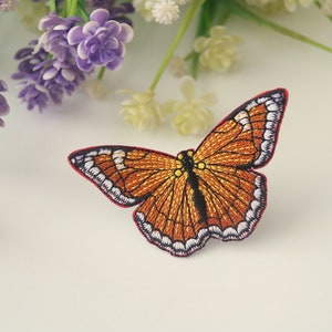 Puede incluir: Un parche de hierro bordado que presenta una mariposa naranja detallada con acentos negros y blancos. La mariposa tiene un contorno rojo y está sobre un fondo blanco.