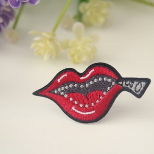 May include: Red embroidered lips with a zipper detail. The zipper is gray with white teeth. The lips are outlined in black.