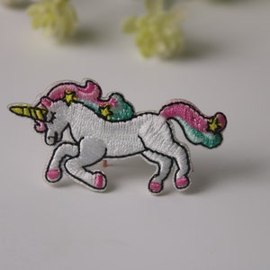 Könnte beinhalten: Weißer gestickter Einhorn-Aufnäher mit rosa und grünem Mähne und Schweif und gelbem Horn. Das Einhorn hat Sterne auf seiner Mähne und seinem Schweif.