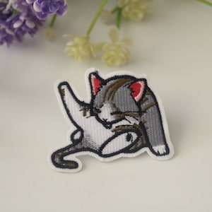 May include: A gray and white embroidered iron-on patch of a cat curled up sleeping. The cat has brown stripes and red ears.