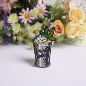 Op de afbeelding: Geborduurde patch met een cocktailglasontwerp met de woorden "mint juice". Het glas is gevuld met een geel en blauw mengsel, bekroond met een groen blad. De patch is zwart en zilver.