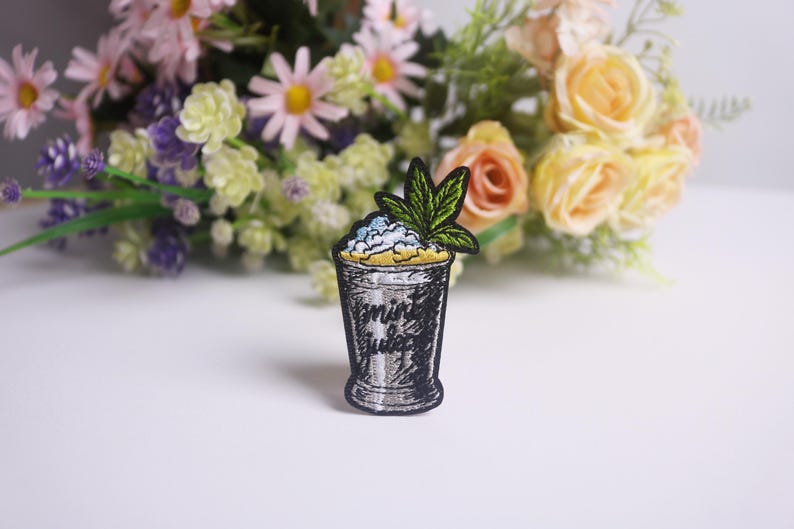 Op de afbeelding: Geborduurde patch met een cocktailglasontwerp met de woorden "mint juice". Het glas is gevuld met een geel en blauw mengsel, bekroond met een groen blad. De patch is zwart en zilver.