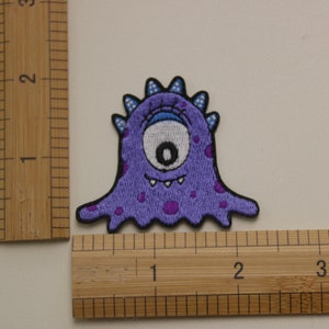 May include: Purple embroidered patch of a one-eyed monster with a toothy grin and purple spots. The monster is about 2 inches tall.