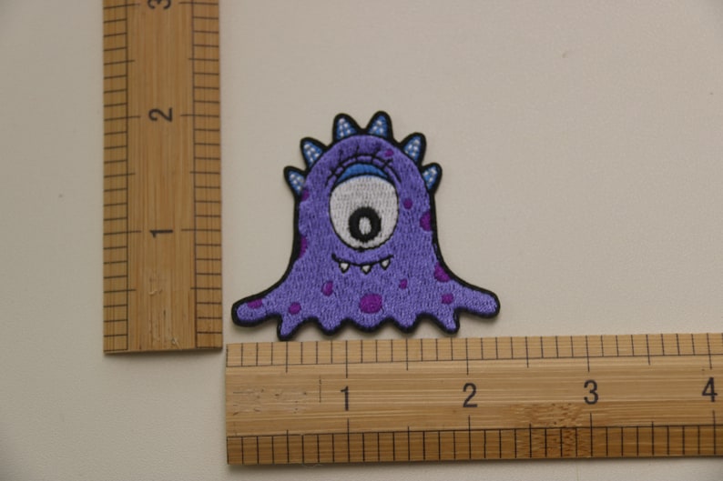 May include: Purple embroidered patch of a one-eyed monster with a toothy grin and purple spots. The monster is about 2 inches tall.