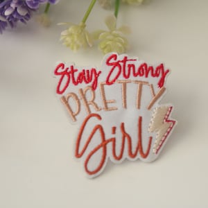May include: White embroidered patch with the text "Stay Strong Pretty Girl" in red and peach thread. A small lightning bolt is embroidered in peach thread to the right of the text.
