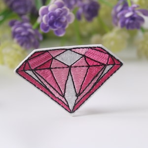 Puede incluir: Un parche bordado en forma de diamante rosa con contornos negros. El parche está sobre un fondo blanco.