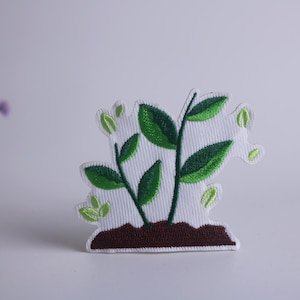 以下が含まれることがあります： 緑の植物のデザインが施された白い刺繍パッチ。植物は複数の葉と茎を持ち、茶色の土壌に根付いています。