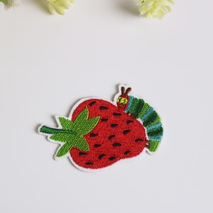 以下が含まれることがあります： 赤いイチゴと緑の茎、緑と青のイモムシが描かれた刺繍パッチ。パッチは白い背景に白い縁取りがされています。このデザインは気まぐれです。