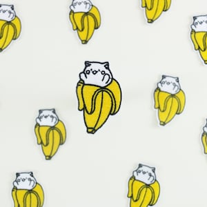 May include: Embroidered patches of a white cat in a yellow banana peel, with black outlines, against a white backdrop. These patches are likely for clothing or accessories. The patches are likely for clothing or accessories.