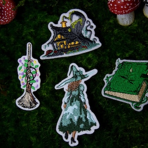 Puede incluir: Parches bordados con temas de brujas. Incluye una casa de brujas, una bruja con sombrero y vestido, una escoba con flores y un libro verde con una libélula. Los parches están sobre un fondo verde y musgoso.