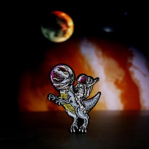 Pode incluir: Um remendo bordado em preto e branco de um dinossauro usando um capacete espacial com um astronauta montando em suas costas. O remendo está sobre um fundo desfocado de um planeta vermelho e laranja.