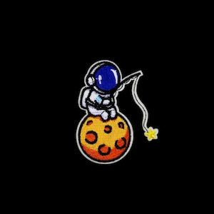 May include: An embroidered patch of an astronaut sitting on a yellow and orange moon, fishing with a line and hook for a yellow star.