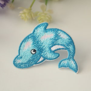 May include: A blue embroidered dolphin patch with white accents and pink spots. The dolphin is jumping out of the water.