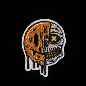 Pode incluir: Patch bordado com um design dividido: metade rosto sorridente, metade caveira. O lado do rosto sorridente é laranja com detalhes gotejantes, o lado da caveira é bege com uma cruz amarela na órbita. Borda branca.