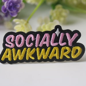 Puede incluir: Parche de planchado negro con el texto "SOCIALLY AWKWARD" bordado en rosa y amarillo.