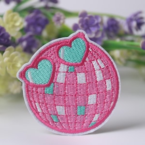 Puede incluir: Bola de disco rosa y blanca con dos corazones turquesa en la parte superior. La bola de disco está bordada con un patrón de cuadros rosa y blanco.