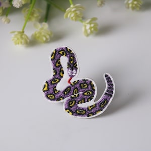 May include: Purple and yellow embroidered snake patch with a black outline. The snake is curled up with its tail in its mouth.