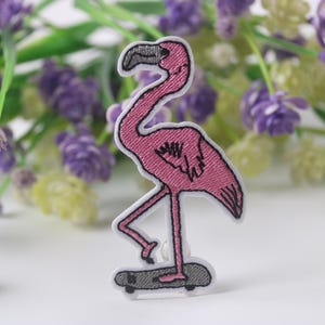 May include: Pink flamingo embroidered patch with black outlines, standing on a skateboard. The flamingo has a long neck and long legs. The skateboard is grey with black wheels.