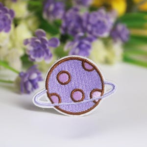 May include: Purple embroidered iron-on patch of a planet with a ring around it. The planet has brown spots.