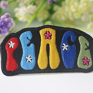 May include: A black fabric patch with the word "PEACE" embroidered in colorful letters. Each letter is outlined in black and has a flower embroidered on it. The flowers are in different colors, including red, pink, and white.