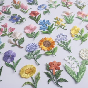 Op de afbeelding: Een verzameling geborduurde bloemvlekken in verschillende kleuren, waaronder blauw, roze, geel en oranje. Elke patch heeft gedetailleerde stiksels en groene bladeren, met een verscheidenheid aan bloemmotieven.