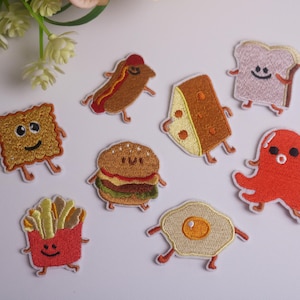 Peut inclure: Patchs brodés représentant des aliments souriants : hot-dog, hamburger, fromage, pain grillé, poulpe, cracker, frites et œuf au plat. Chaque patch a des bras et des jambes. Les patchs sont colorés et fantaisistes.