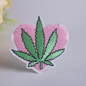 Könnte beinhalten: Ein rosafarbenes herzförmiges Patch mit einem gestickten grünen Marihuana-Blatt-Design.