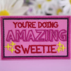 Könnte beinhalten: Ein pinkfarbener, gestickter Aufnäher mit den Worten "YOU'RE DOING AMAZING SWEETIE" in rotem und pinkfarbenem Garn. Der Aufnäher hat einen schwarzen Rand und gelbe Sternakzente, ideal zum Aufnähen auf Kleidung oder Accessoires.