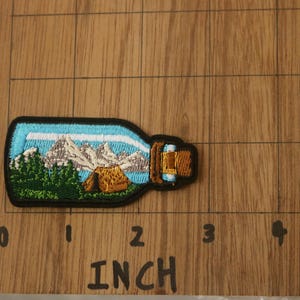 May include: An embroidered patch depicting a mountain scene with a small cabin in the foreground. The patch is shaped like a bottle with a cork top, and is blue and white.