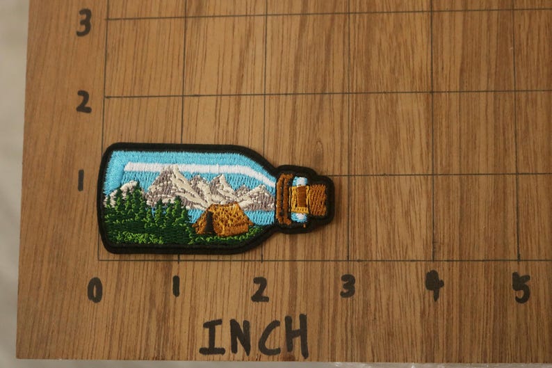 May include: An embroidered patch depicting a mountain scene with a small cabin in the foreground. The patch is shaped like a bottle with a cork top, and is blue and white.