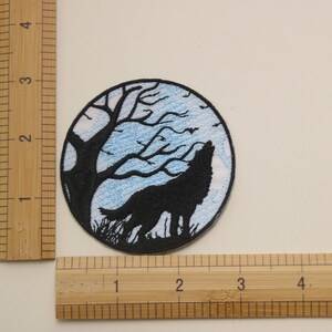 Op de afbeelding: Gestikt patch met een zwart silhouet van een wolf die huilt naar de maan. De maan is blauw met witte wolken en een zwarte boom op de achtergrond.