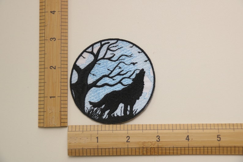 Op de afbeelding: Gestikt patch met een zwart silhouet van een wolf die huilt naar de maan. De maan is blauw met witte wolken en een zwarte boom op de achtergrond.