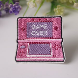 Könnte beinhalten: Rosa und lila gestickter Bügelaufkleber einer tragbaren Videospielkonsole mit dem Text "GAME OVER" auf dem Bildschirm.