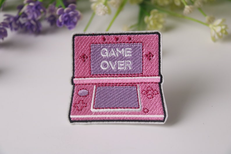 Może przedstawiać: Haftowany naszywka do prasowania w kolorze r&oacute;żowym i fioletowym przedstawiająca przenośną konsolę do gier wideo z wyświetlonym na ekranie napisem "GAME OVER".
