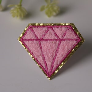 May include: Pink diamond-shaped patch with a gold glitter border. The patch is made of a fuzzy fabric.
