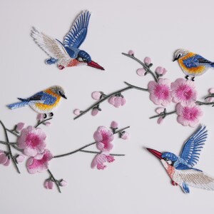May include: Embroidered patches featuring colorful birds with blue, yellow, and white feathers, perched on branches with pink cherry blossoms. The patches are detailed and suitable for clothing or craft projects.