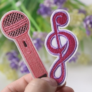Könnte beinhalten: Zwei gestickte Bügelbilder, ein rosa Mikrofon und ein violetter Violinschlüssel, auf weißem Hintergrund.