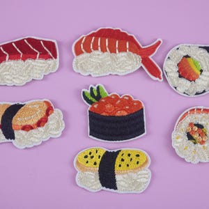 Puede incluir: Parches de sushi bordados en varios estilos, incluyendo nigiri y rollos maki, con detalles en rojo, naranja y amarillo. Los parches están sobre un fondo lila claro, mostrando la costura detallada y el diseño.